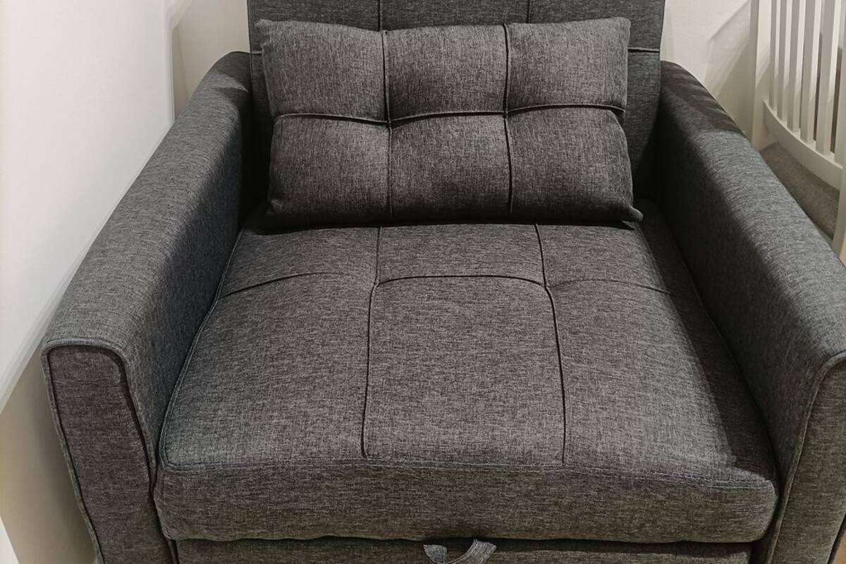Harmaa, kangasverhoiltu nojatuoli, jossa on tyyny, ja se on sijoitettu valkoista seinää vasten.