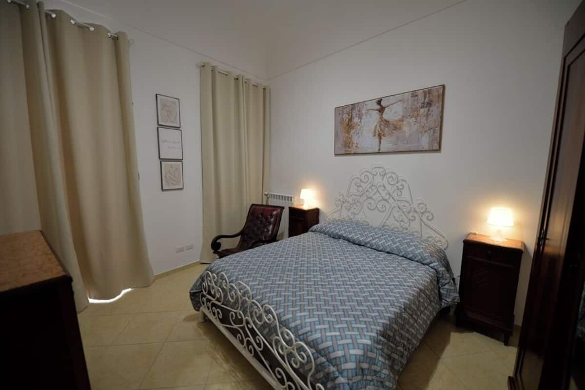 Ein Schlafzimmer mit Bett, Vorhängen, einem Sessel, einem Nachttisch und Wandbildern.