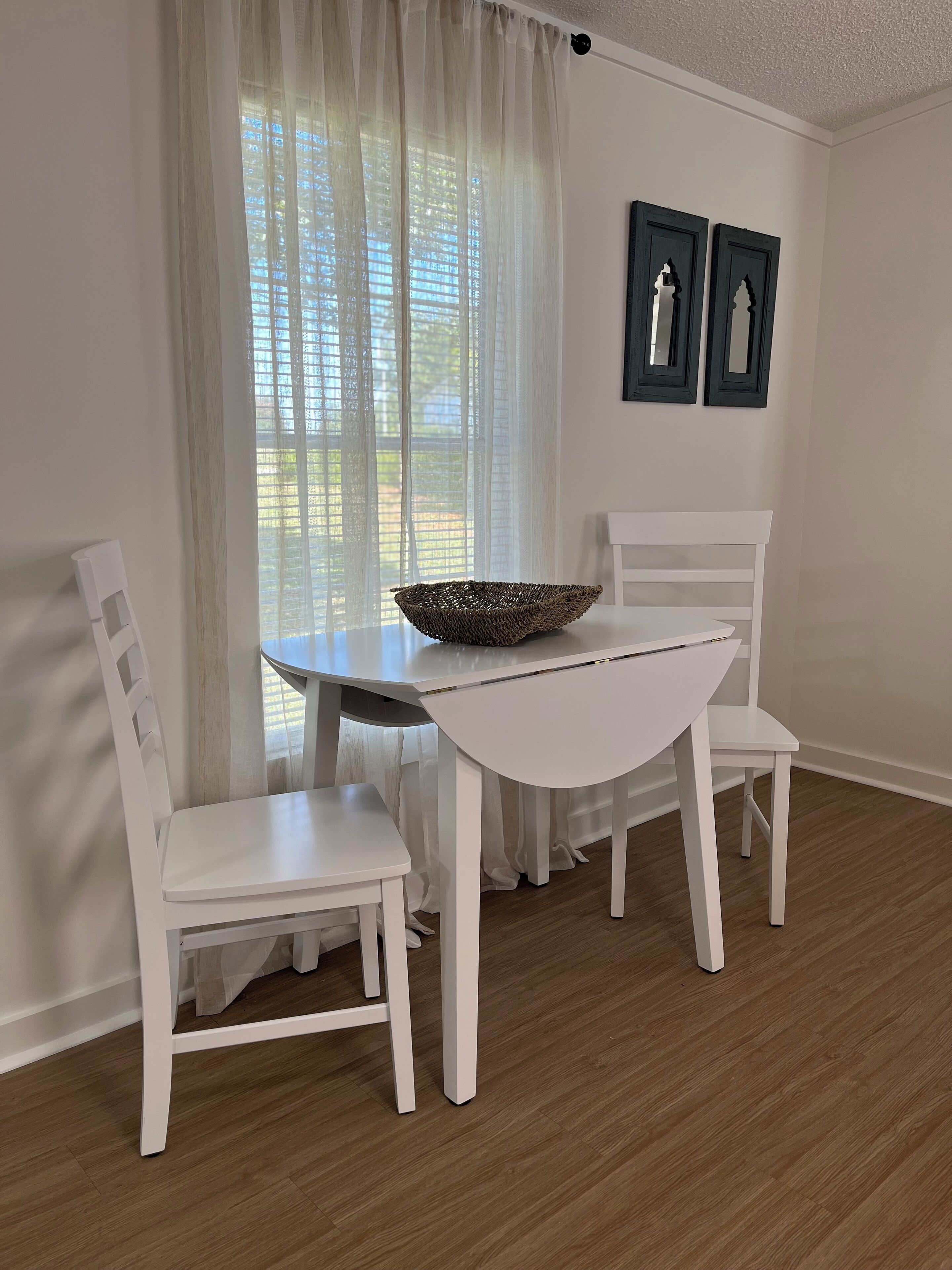 Een eethoek met een witte tafel en stoelen, een geweven mand op tafel, en een raam met transparante gordijnen.