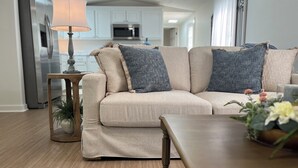 Ein Wohnzimmer mit einem beigefarbenen Sofa, gemusterten Kissen, einem Holznachttisch und einer Lampe.