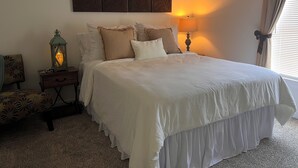 Ein ordentlich bezogenes Bett mit weißer Daunendecke und mehreren Kissen, ein Nachttisch mit Lampe, eine Zierleiste an der Wand und ein Fenster mit Vorhängen.