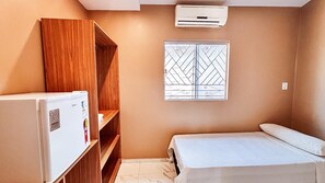 A small, compact room with a bed, a wooden wardrobe, a white refrigerator, and a window with a metal grille. - HOTEL AREJAR (Juruti)