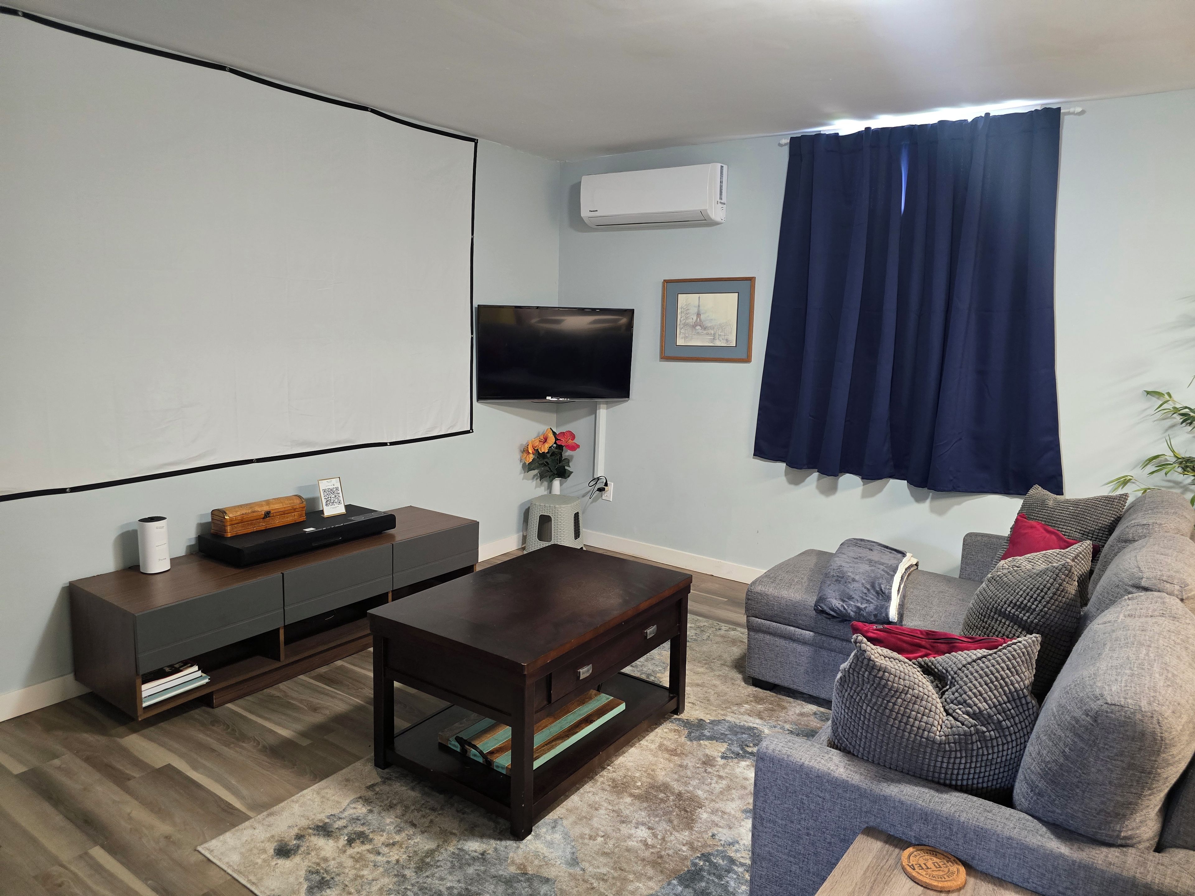 Una sala de estar con televisor de pantalla plana, una mesa de centro de madera oscura, un sofá gris con cojines estampados y una cortina azul.