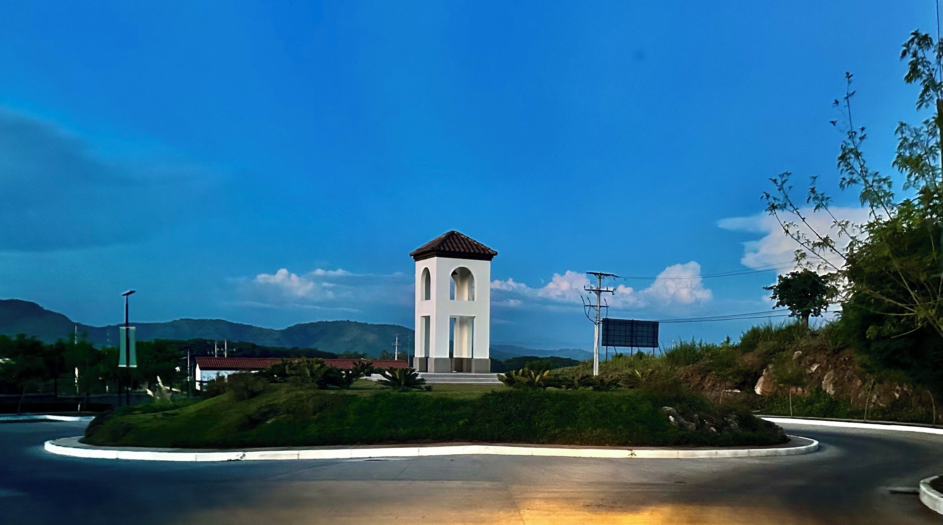 A white tower with a red roof, surrounded by greenery, at a roundabout with a clear sky.