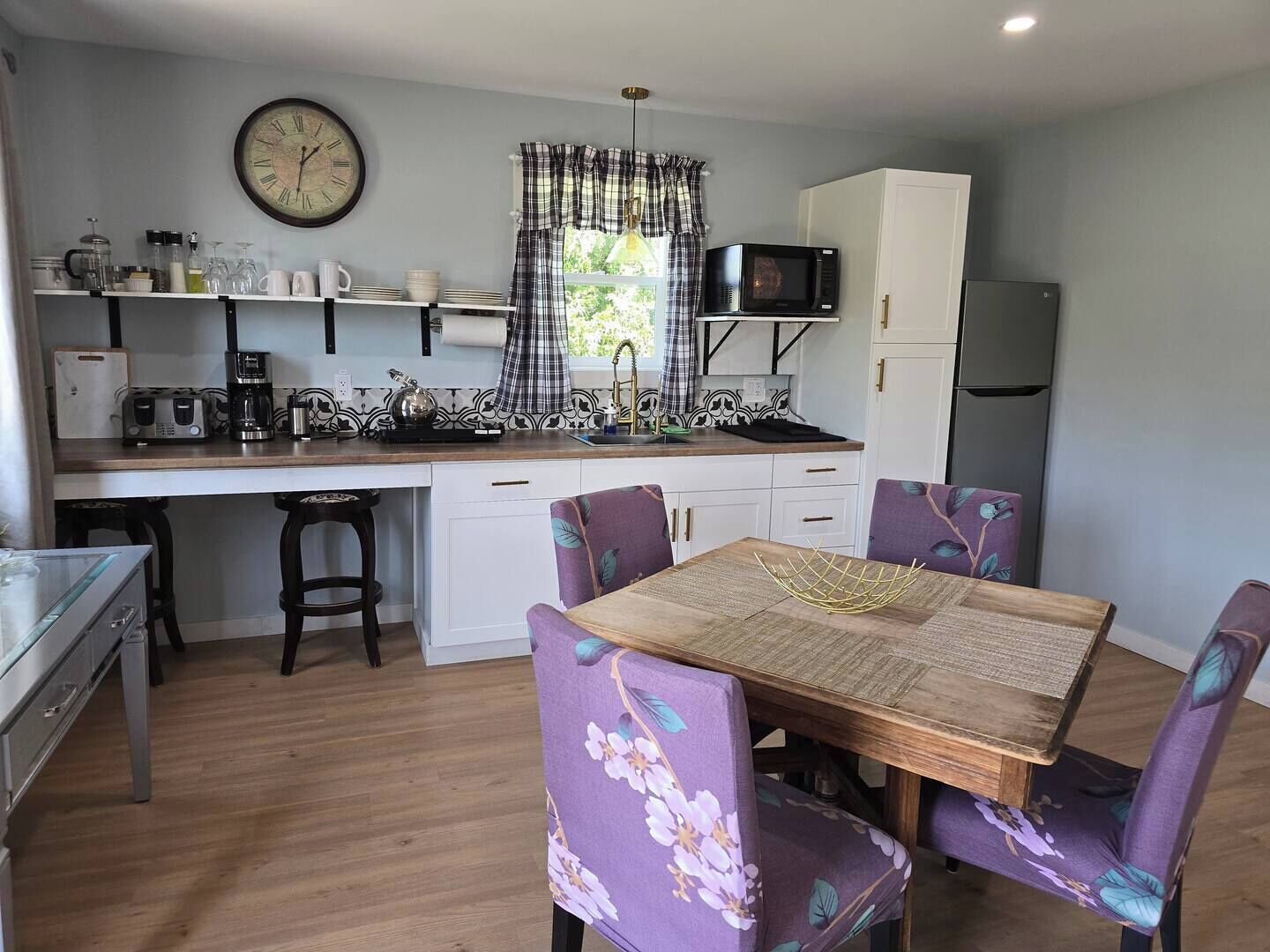 Een compacte keuken met een houten tafel en paarse stoelen met bloemenprint, een klok aan de muur en een magnetron boven het aanrecht.