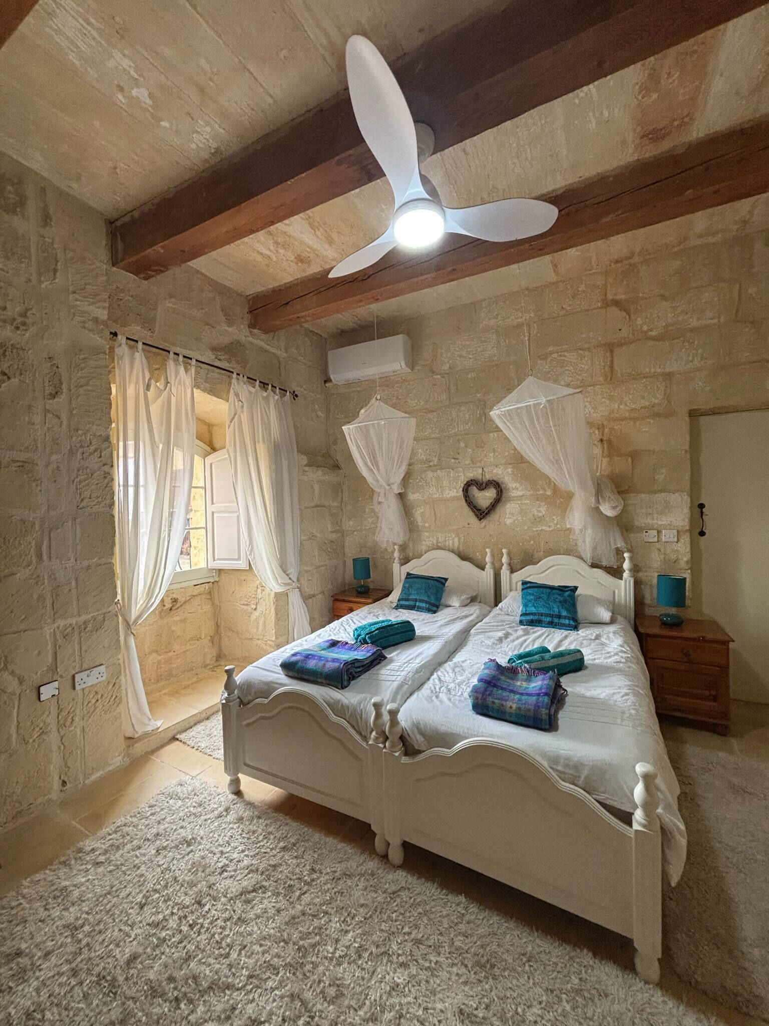 Une chambre avec un lit blanc, deux tables de chevet, un ventilateur de plafond et un mur en pierre.