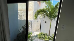 A view through a window of a courtyard with a palm tree, potted plants, and a brick pathway. - Lavish Studio w Kitchen Downtown (Isla Mujeres)