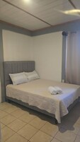 Una cama con cabecera gris, una colcha blanca y dos toallas enrolladas sobre la cama.
