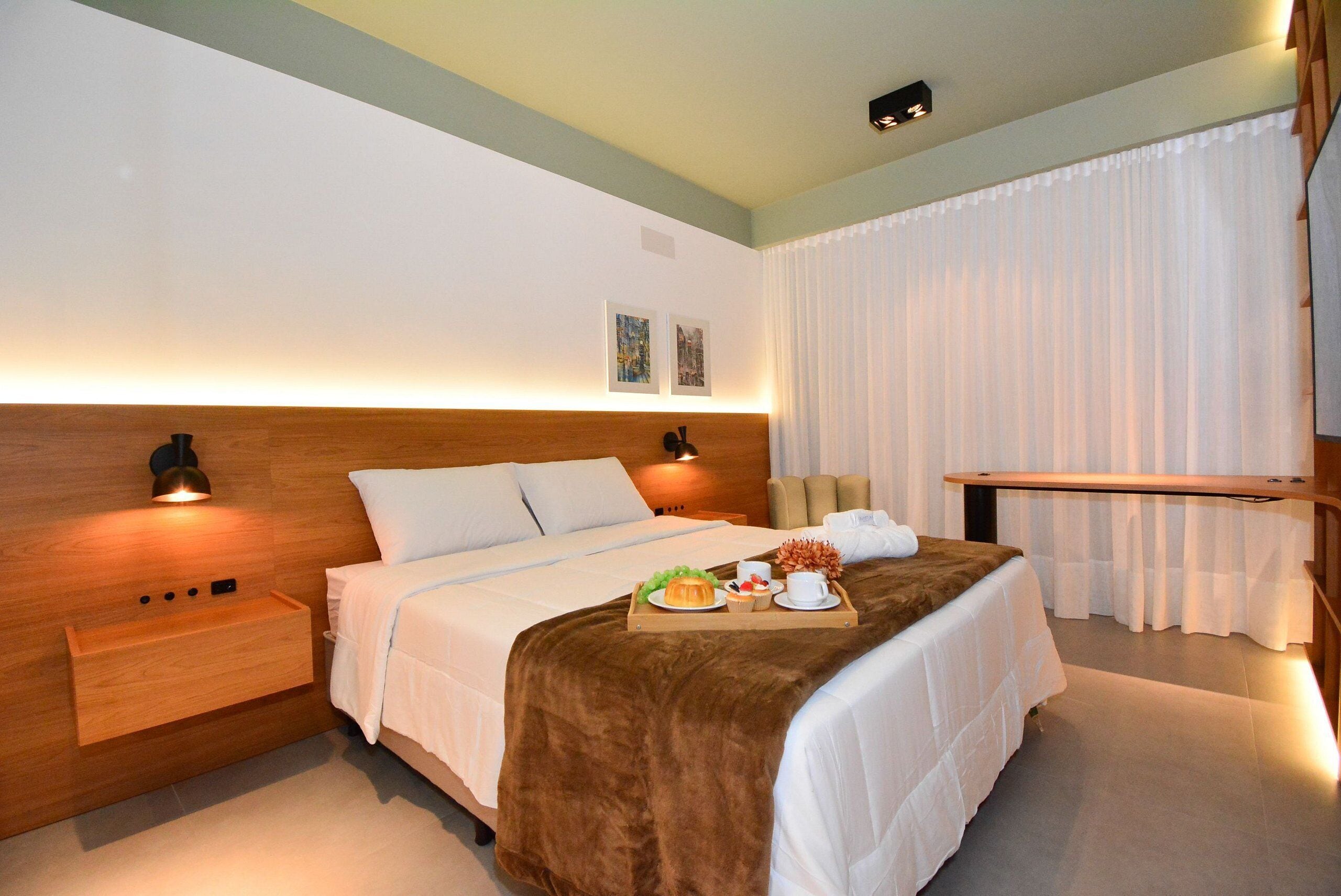 Una habitación de hotel moderna con cabecera de madera, una mesita de noche con lámpara, cama con manta marrón y una bandeja con artículos para el desayuno.