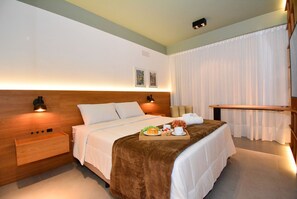 Una habitación de hotel moderna con cabecera de madera, una mesita de noche con lámpara, cama con manta marrón y una bandeja con artículos para el desayuno.