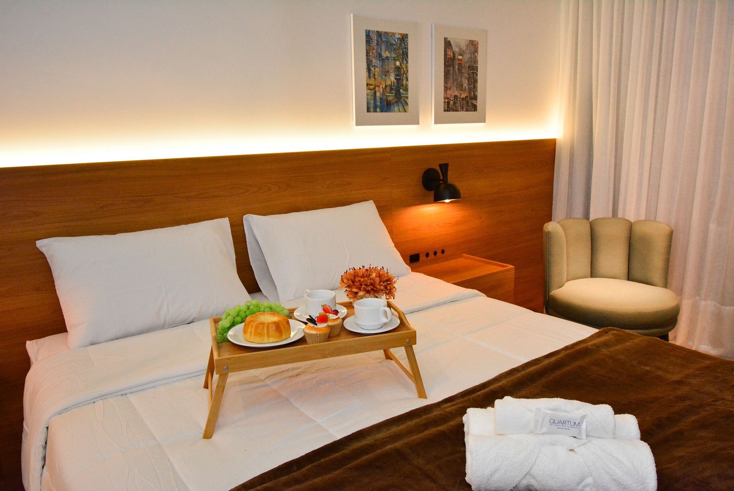 A hotel room with a bed, a bedside table with a lamp, a chair, and a tray with food and drinks.