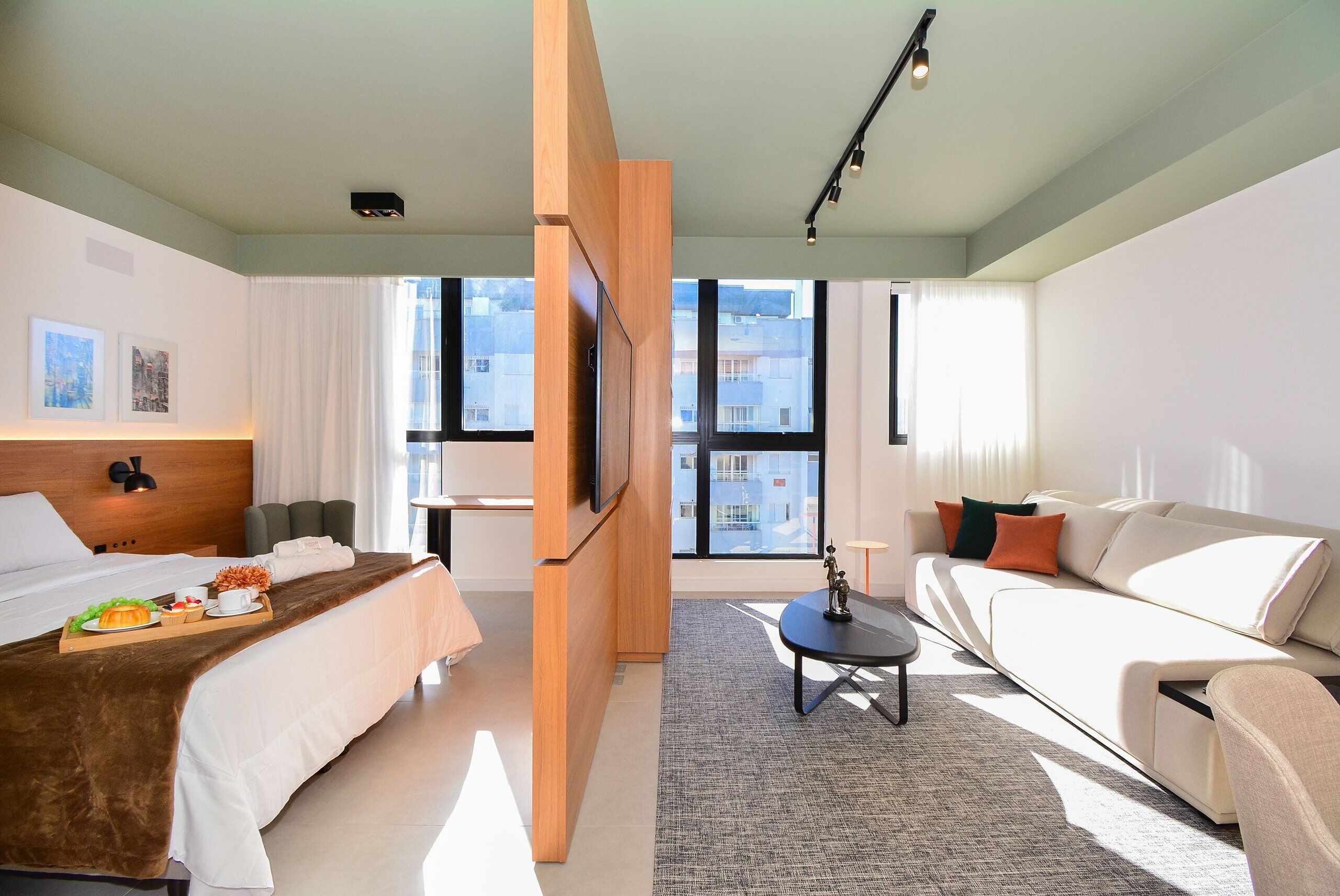 A modern hotel room with a bed, sofa, TV, and large windows offering city views.
