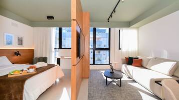 Ein modernes Hotelzimmer mit Bett, Sofa, Fernseher und großen Fenstern mit Blick auf die Stadt.