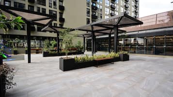 An outdoor courtyard with modern architectural design, featuring geometric patterns on the floor, black planters with greenery, and a covered seating area.