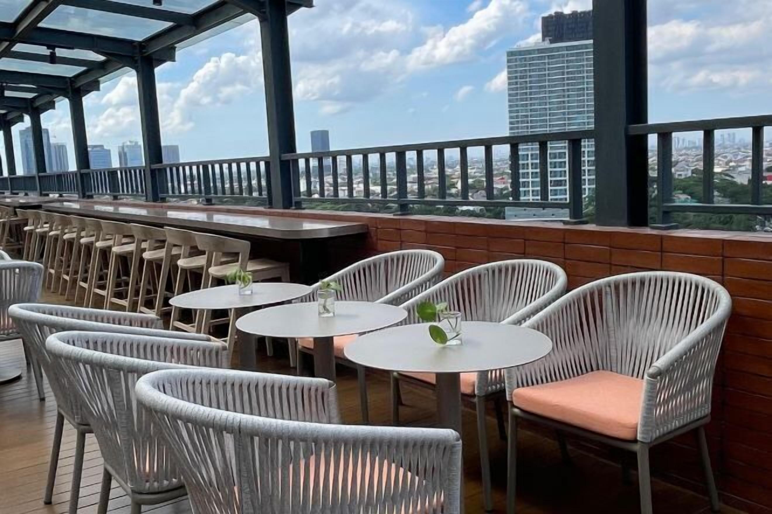 An outdoor seating area with wicker chairs and round tables, overlooking a cityscape.