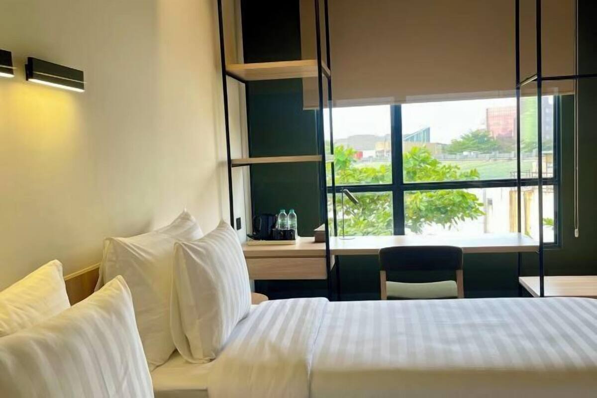 Habitación de hotel con cama, escritorio, silla y ventana con vistas a zonas verdes y edificios.