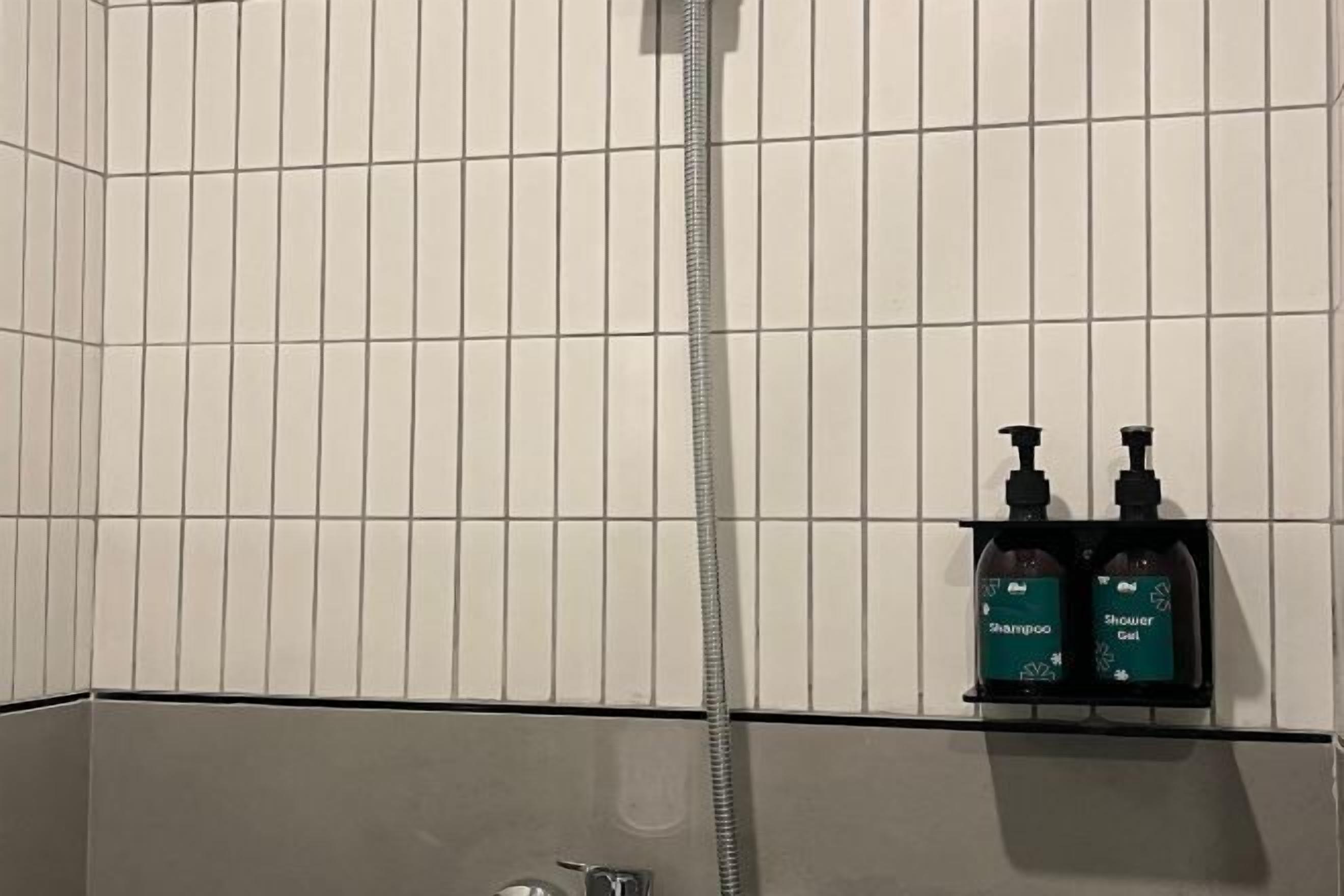 A bathroom with white tiled walls, a shower, and a shelf with two bottles of shampoo and shower gel.