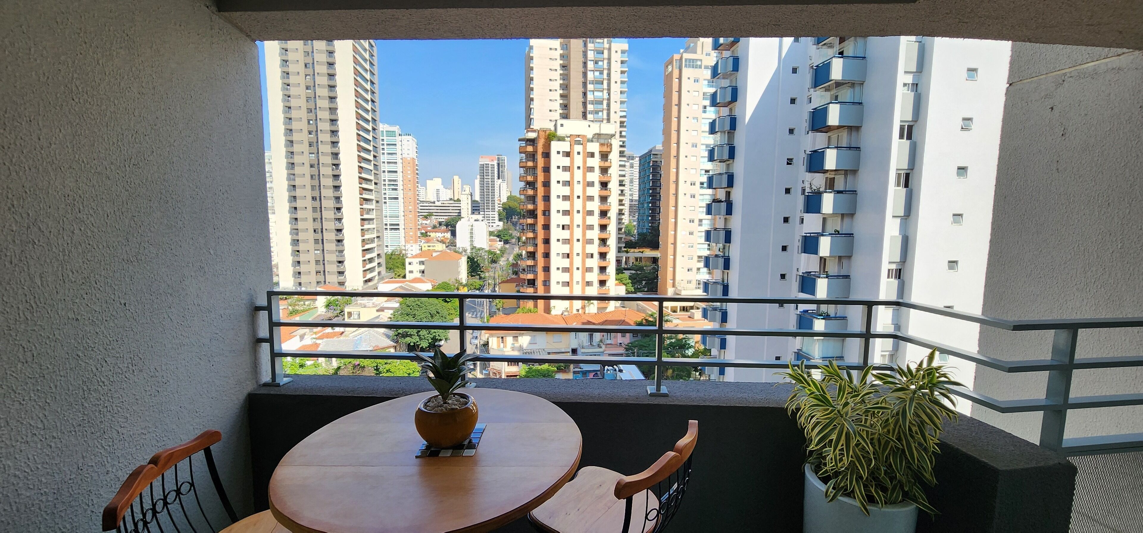 Uma varanda com uma mesa redonda e cadeiras, um vaso com planta e vista para edifícios altos.
