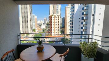 Uma varanda com uma mesa redonda e cadeiras, um vaso com planta e vista para edifícios altos.