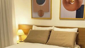 A neatly made bed with a beige comforter and pillows, a bedside lamp, and two abstract paintings on the wall. - Comfortable studio apartment in Perdizes - Allianz Parque with pool and rooftop. (São Paulo)