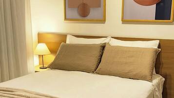 Cama bem arrumada com edredom bege e almofadas, luminária de cabeceira e duas pinturas abstratas na parede.