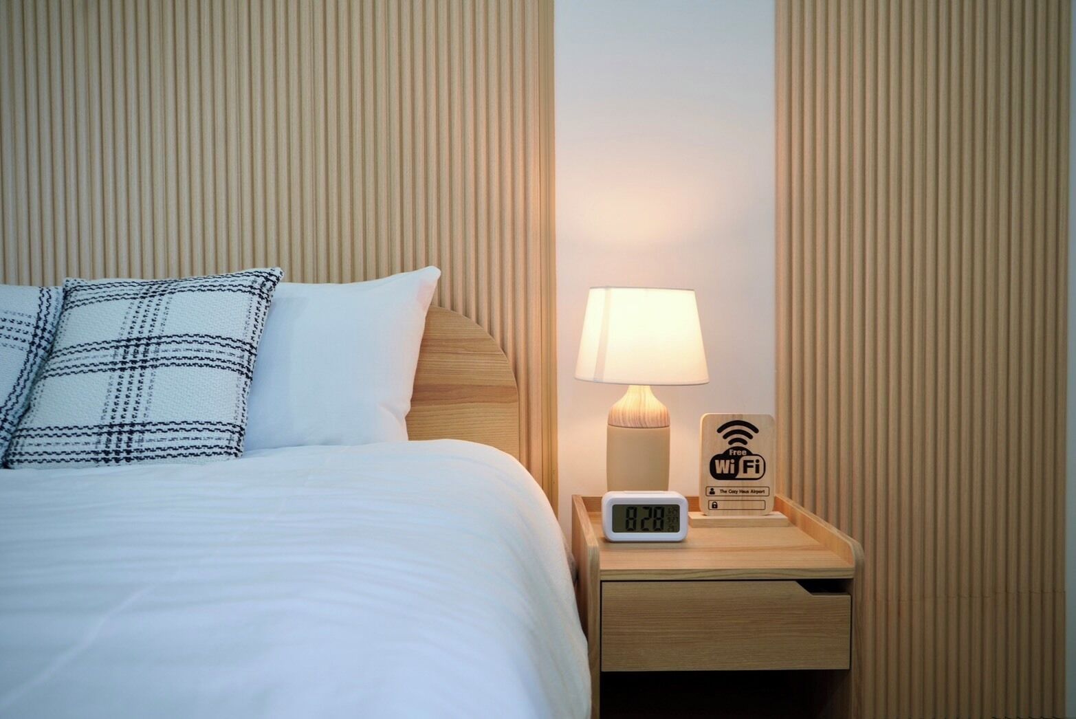 A neatly made bed with white bedding and a plaid pillow, a bedside lamp, a digital clock, and a Wi-Fi sign on a wooden nightstand.