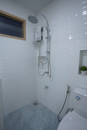 A bathroom with white tiled walls, a shower with a handheld showerhead, a wall-mounted faucet, and a toilet. - The Cozy Haus Airport (Mai Khao)