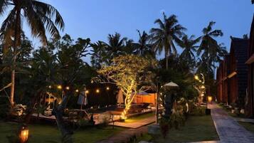 Un resort tropical con un camino bien iluminado, palmeras y un edificio con techo de paja.