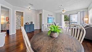 A spacious living room with a wooden dining table, white chairs, and a large window offering a view of the outdoors. - Wolf Bay Villas 208: Waterfront (Orange Beach)