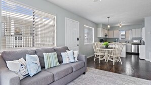 A modern living room with a grey sofa, white dining table, and chairs, a kitchen in the background, and large windows with blinds. - Wolf Bay Villas 208: Waterfront (Orange Beach)