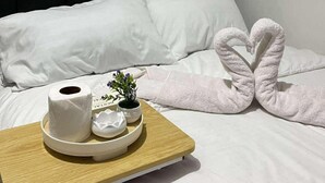 Siististi pedattu sänky valkoisilla lakanoilla, sydämenmuotoinen pyyhe, pieni tarjoilualusta, jossa on hygieniatuotteita, ja pieni kasvi.