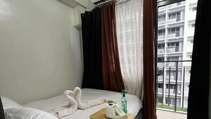 Hotellihuone, jossa on sänky, yöpöytä, jossa on hygieniatarvikkeita, ikkuna, jossa on verhot, ja näkymä rakennukseen.