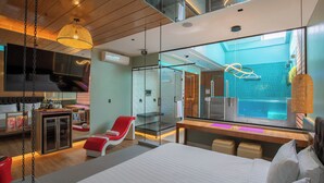 A modern hotel room with a large bed, a seating area with a red chair, a glass-enclosed shower, and a view of a pool. - Liv Motel Ecuador (Quito)