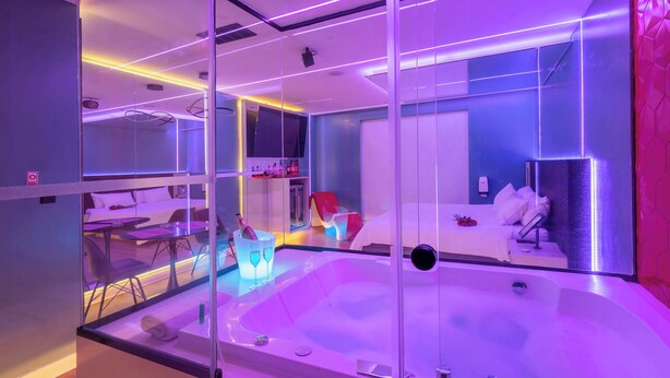 A modern hotel room with a glass-enclosed jacuzzi, a bed, a small table with a chair, and a seating area with a red chair. - Liv Motel Ecuador (Quito)