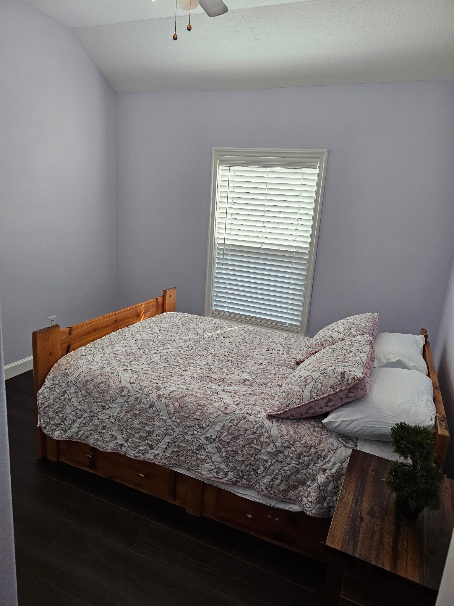 A bedroom with a wooden bed frame, a floral bedspread, and a window with blinds.