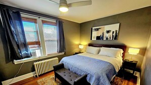 Een slaapkamer met een bed, bedlampen, een radiator, een raam met gordijnen, een plafondventilator, een vloerkleed en een bankje.