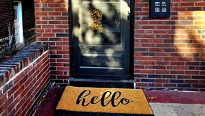 Een zwarte deur met een 'hello' mat ervoor, een bakstenen muur en een brievenbus aan de rechterkant.