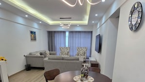 A modern living room with a dining area, featuring a round table with chairs, a sofa set, a flat-screen TV, and a decorative wall clock. - Marina Bay Suites (Nairobi)