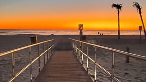 Een houten looppad dat naar een strand leidt, met palmbomen en een zonsondergang op de achtergrond.