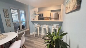 Een kleine keuken met witte kastjes, een magnetron en een koelkast. Drie witte barkrukken en een eettafel met stoelen.