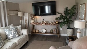 Un soggiorno con divano, tavolino, TV a parete, mensola decorativa con piante e candele, e una pianta in vaso.