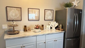 Una cucina con piano di lavoro in marmo, mobili bianchi e frigorifero in acciaio inossidabile. Si vedono tre quadri appesi al muro, una pianta sul piano di lavoro e diversi oggetti da cucina.