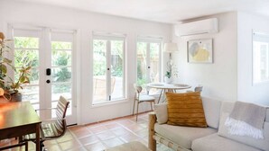 A bright, open living area with a sofa, a dining table, and chairs. There are large windows providing natural light and views of the outdoors. - Montecito Orchard Cottage (Montecito)