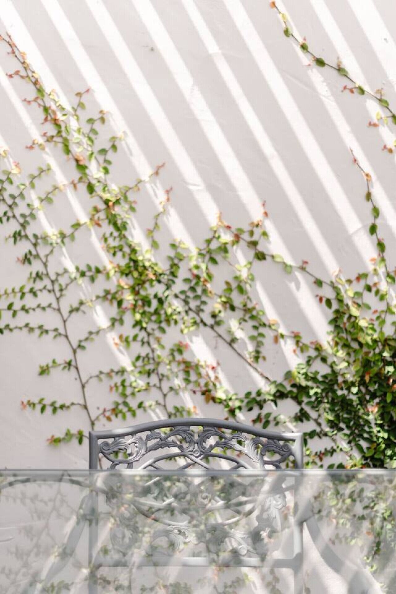 A table with a glass top and metal frame, positioned in front of a white wall with vertical shadows.