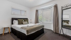 A bedroom with a bed, bedside table, window with blinds, and a mirror. - Bayfair Break - Mt Maunganui (Mt Maunganui)