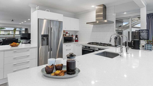 A modern kitchen with white cabinetry, stainless steel appliances, and a central island with a coffee pot and pastries. - Bayfair Break - Mt Maunganui (Mt Maunganui)