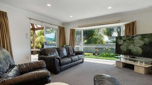 A modern living room with a leather sofa, a flat-screen TV, and a large window offering a view of the outdoors. - Bayfair Break - Mt Maunganui (Mt Maunganui)