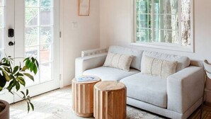 Una sala de estar moderna con un sofá blanco, dos mesas auxiliares de madera, un ventanal grande y un techo con vigas a la vista.