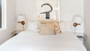 Un dormitorio con una cama grande, dos mesitas de noche con lámparas, una obra de arte enmarcada sobre la cama y una ventana a la izquierda.