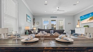 A dining area with a wooden table set for two, a ceiling fan, and a view of the living room with a couch and a sign reading "BEACH HOUSE". - New! Steps to Beach & Lagoon~Pool~Family Fun~Paddle Boards (Gulf Shores)